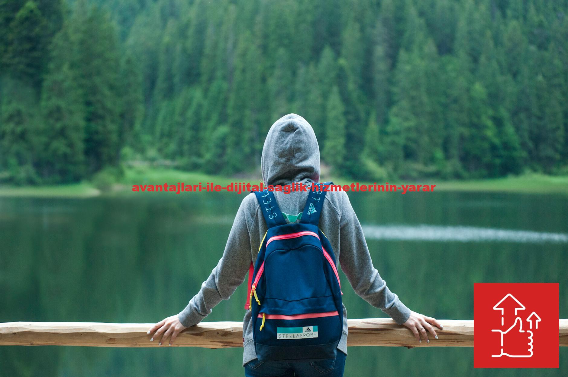 A person in a hoodie stands by a serene lake surrounded by lush greenery, enjoying the peaceful outdoors.
